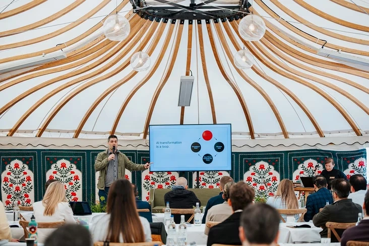 Philip Lakin of Zapier speaking into a microphone beside a screen reading AI transformation is a loop, in front of seated attendees in a tent